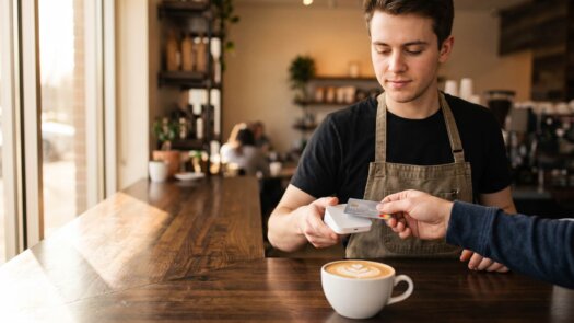 Barista hält einer Kundin den kleinen Kartenleser hin, Cappuccino auf dem Tresen